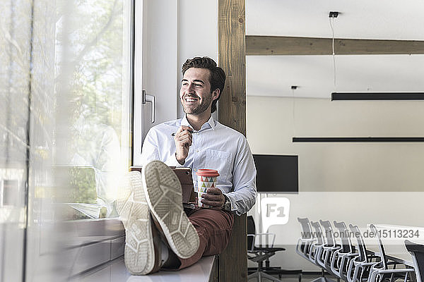 Young businessman sitting on windowsill  using digital tablet  drinking coffee