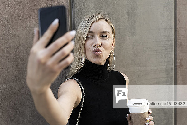 Portrait of blond woman with coffee to go taking selfie with smartphone