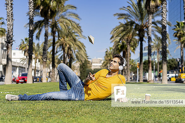 Spain  Barcelona  man lying on lawn in the city with headphones throwing up notebook