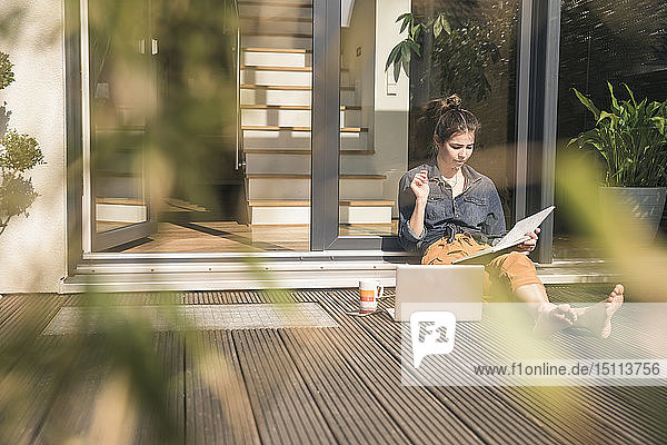 Junge Frau sitzt zu Hause auf der Terrasse und arbeitet mit Buch und Laptop