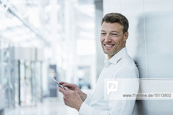 Porträt eines lächelnden Geschäftsmannes  der sich an eine Wand lehnt und ein Mobiltelefon hält