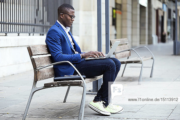 Young businessman wearing blue suit jacket sitting on bench and using laptop
