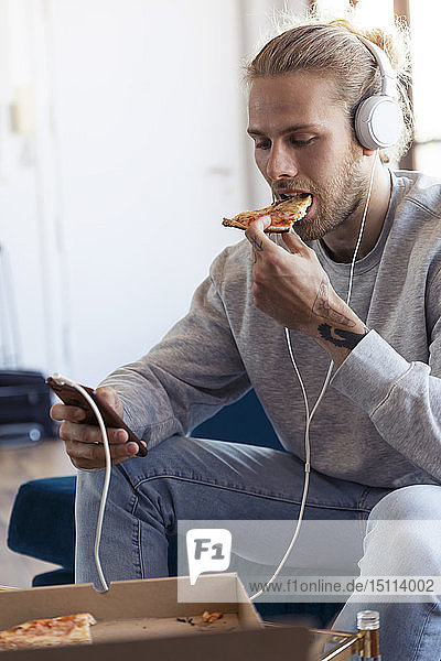 Junger Mann auf Couch mit Handy und Kopfhörer beim Pizzaessen