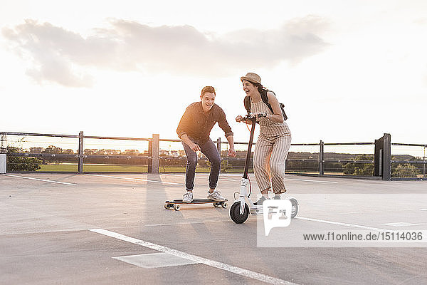 Young man and woman riding on longboard and electric scooter on parking deck at sunset