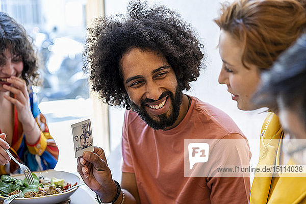 Glücklicher Mann zeigt Freunden beim Mittagessen in einem Restaurant ein Sofortbild