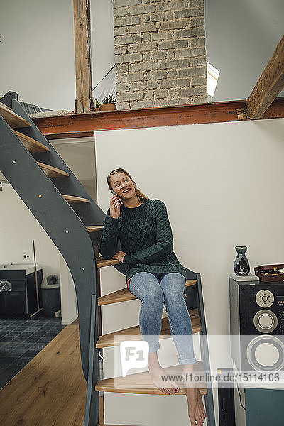 Lächelnde junge Frau sitzt zu Hause auf der Treppe und telefoniert per Handy