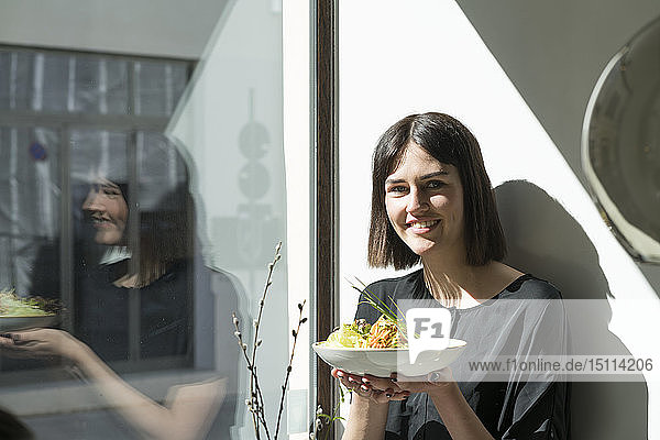 Lächelnde junge Frau steht bei Sonnenschein am Fenster und hält einen Teller Salat
