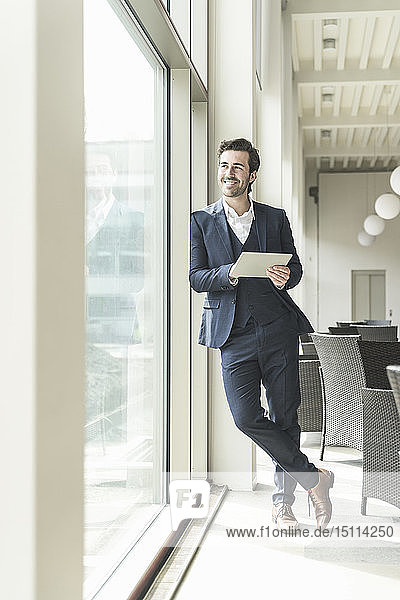 Young businessman standing in office building  using digital tablet  looking out of window