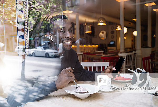 Porträt eines lächelnden jungen Mannes mit Kopfhörern und Smartphone hinter einer Fensterscheibe in einem Restaurant