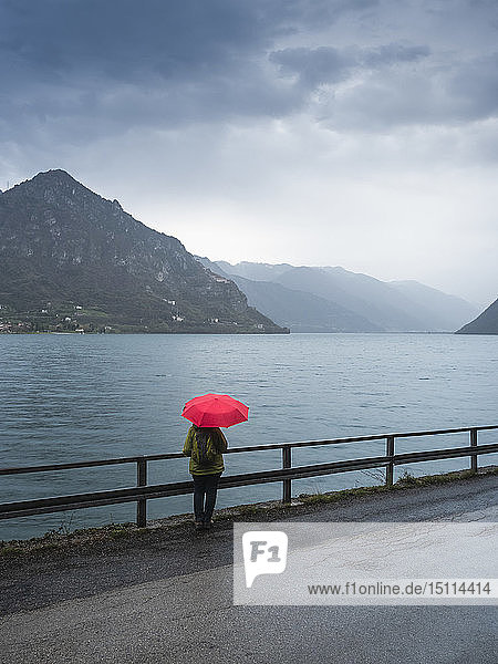 Italy  Lombardy  back view of senior woman with backpack and red umbrella looking at Lake Idro