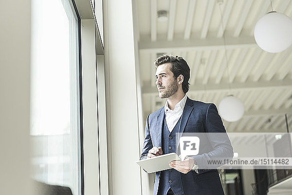Erfolgreicher Manager  der in einem modernen Bürogebäude steht  einen Laptop benutzt und aus dem Fenster schaut