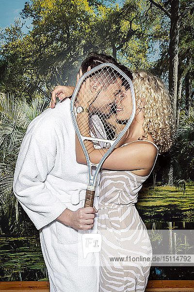 Junges Paar steht vor einer Fototapete  küsst sich hinter einem Tennisschläger