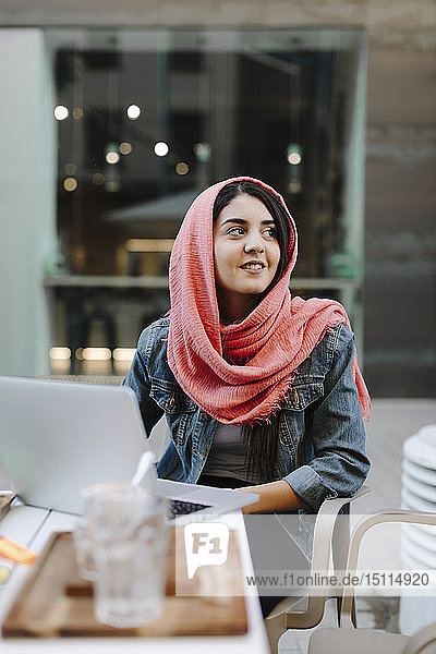 Lächelnde junge Frau mit Laptop und Kopftuch in einem Straßencafé