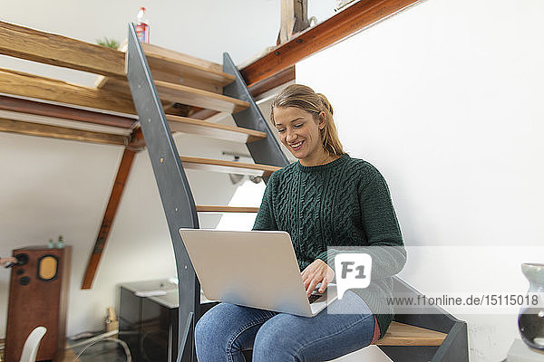 Lächelnde junge Frau sitzt zu Hause auf der Treppe und benutzt ihren Laptop