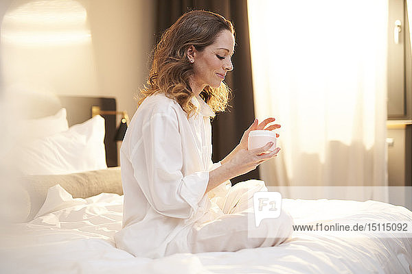 Brünette Frau sitzt auf dem Bett und trägt Schönheitscreme auf