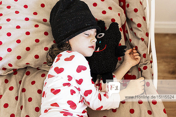 Kleines Mädchen mit Mütze schläft mit ihrem Stofftier im Bett