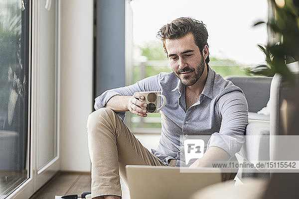 Junger Mann sitzt zu Hause auf dem Boden  benutzt Laptop  trinkt Kaffee