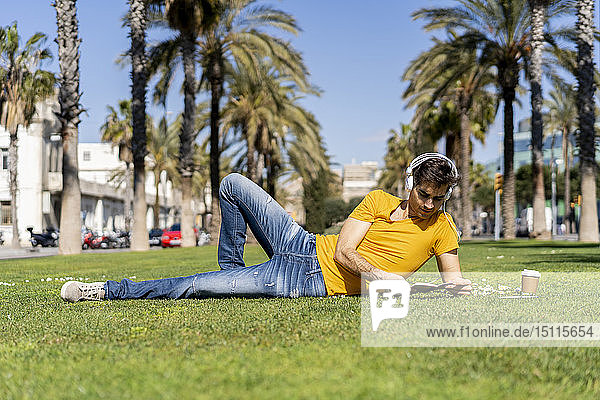 Spanien  Barcelona  auf dem Rasen liegender Mann in der Stadt mit Kopfhörern und Notebook