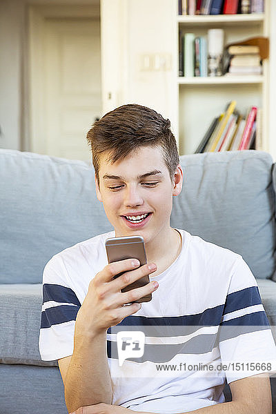 Porträt eines lächelnden Teenagers  der mit seinem Handy vor der Couch sitzt