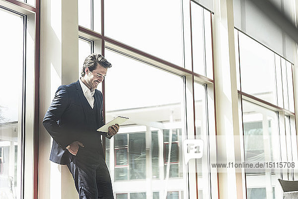 Young businessman standing in office building  using digital tablet  leaning on window