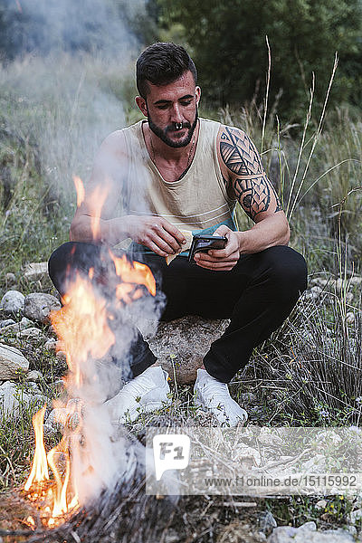 Mann sitzt mit Handy am Lagerfeuer in ländlicher Landschaft