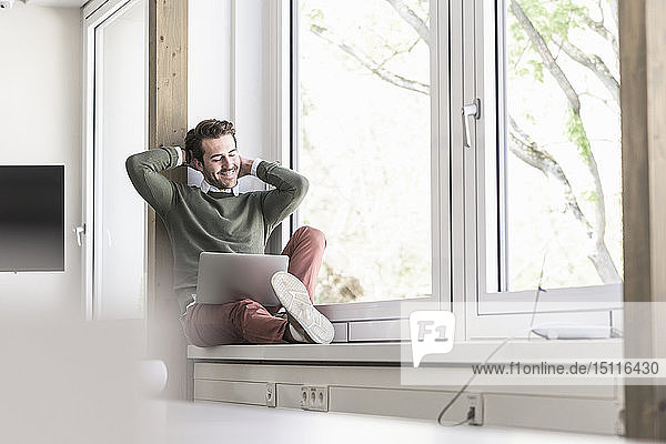Junger Geschäftsmann sitzt auf der Fensterbank und benutzt einen Laptop