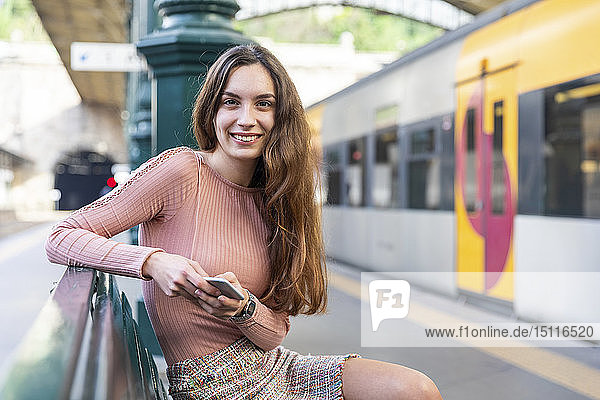 Portrait of smiling young woman with smartphone waiting on bench on platform  Porto  Portugal