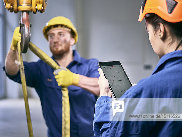 Industriearbeiter fixiert Hebezeuggehänge am Hallenkran  Kollegin steuert mit digitalem Tablett