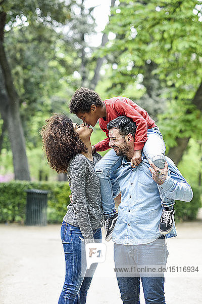 Vater trägt seinen Sohn huckepack in einem Park  während der Sohn seine Mutter küsst