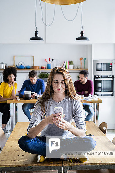 Woman with cell phone sitting on dining table at home with friends in background