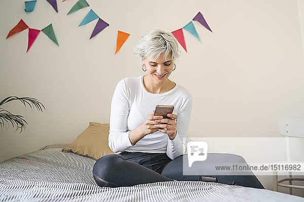 Lächelnde Frau  die zu Hause am Bett ein Mobiltelefon benutzt