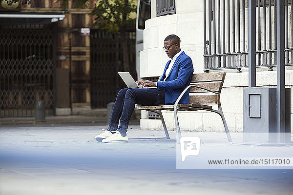 Junger Geschäftsmann in blauer Anzugjacke sitzt auf Bank und benutzt Laptop