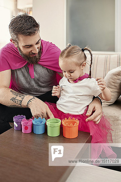 Vater und Tochter im Superhelden- und Superfrau-Kostüm  spielen mit gestapelten Tassen