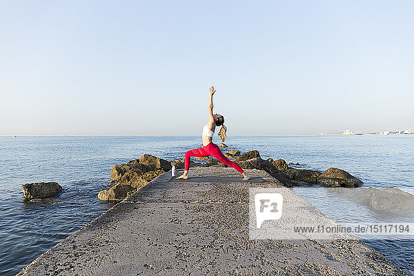 Junge Frau  die am Strand Yoga praktiziert und dabei eine Kriegerpose einnimmt
