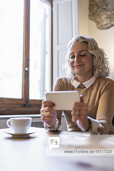 Porträt einer lächelnden reifen Geschäftsfrau  die mit einer Tasse Kaffee am Tisch sitzt und auf ihr Handy schaut