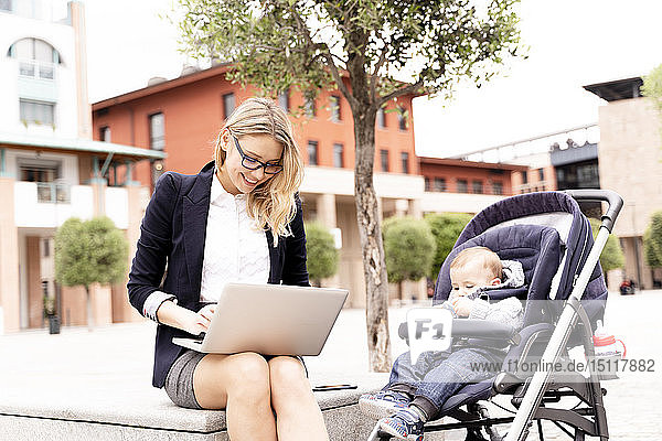 Junge Geschäftsfrau mit einem kleinen Jungen im Kinderwagen  der im Freien am Laptop arbeitet