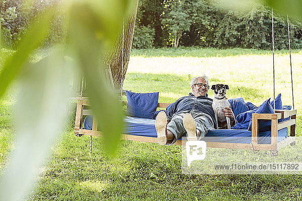 Älterer Mann entspannt sich auf einem Schaukelbett in seinem Garten mit seinem Hund
