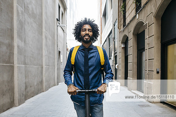 Porträt eines lächelnden jungen Mannes mit Rucksack auf einem E-Scooter nach der Arbeit  Barcelona  Spanien