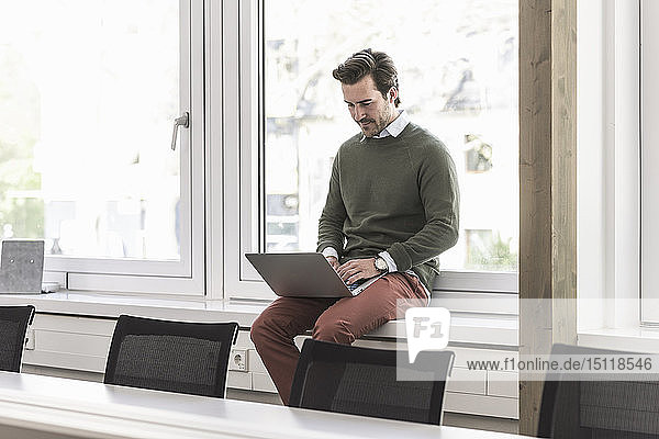Junger Geschäftsmann sitzt auf der Fensterbank und benutzt einen Laptop