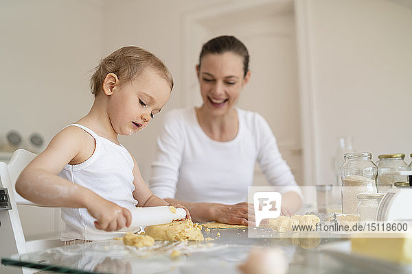 Mother and little daughter with dough roll making a cake together in kitchen at home