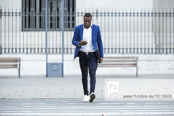 Junger Geschäftsmann in blauer Anzugjacke überquert die Straße und benutzt ein Smartphone