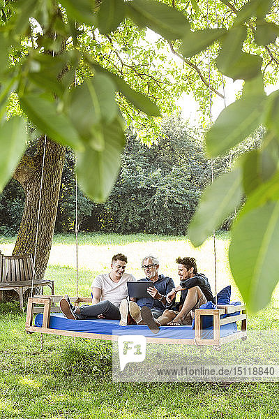 Männer einer Familie sitzen auf einem Schaukelbett im Garten  unterhalten sich  benutzen ein digitales Tablet