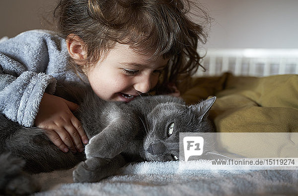 Happy toddler girl cuddling grey cat lying on bed