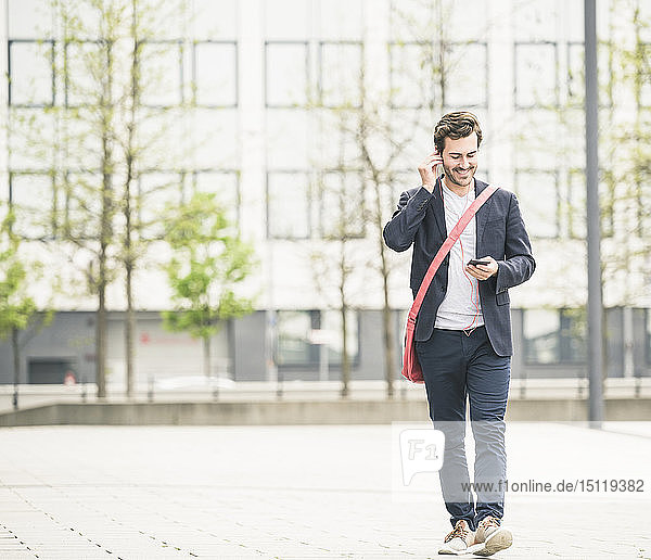 Smiling businessman walking in the city with cell phone and earphones