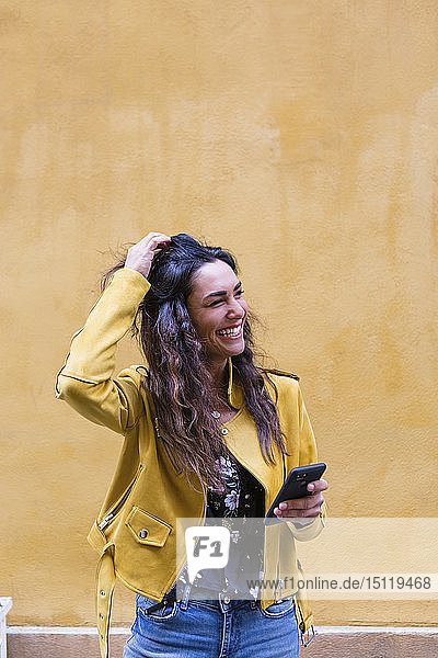 Porträt einer jungen lachenden Frau  die eine gelbe Lederjacke trägt und ein Smartphone in der Hand hält