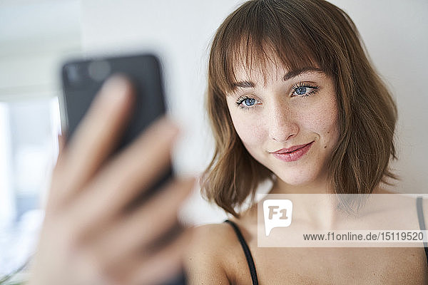 Porträt einer lächelnden jungen Frau  die sich zu Hause mit einem Smartphone selbstständig macht