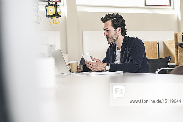 Junger Geschäftsmann sitzt im Büro und benutzt Laptop und Smartphone