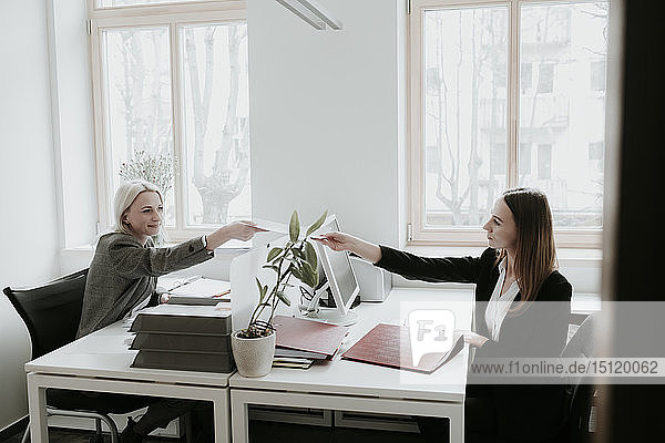 Two young women working at desk in office handing over paper