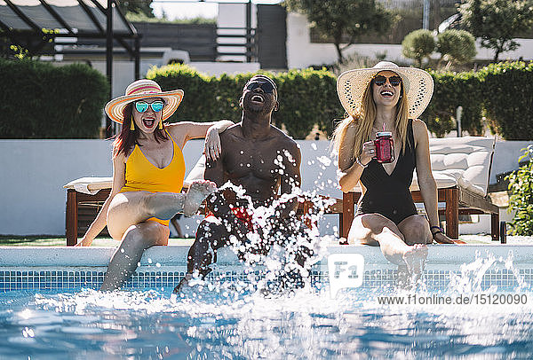 Zwei glückliche Frauen und ein Mann sitzen am Pool und spritzen Wasser