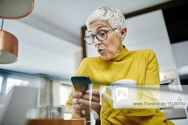 Ältere Frau trinkt Kaffee und überprüft Textnachrichten auf ihrem Smartphone